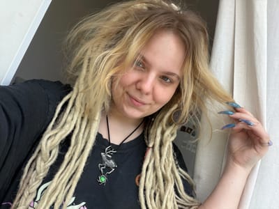 The image shows a young person with blonde dreadlocks, wearing a black t-shirt and a necklace with a deer pendant. The person is looking at the camera and touching their hair. The background is a light-colored curtain and a white wall. The scene appears to be indoors. No specific details about Vostochny District, Russia are visible or inferable from the image.