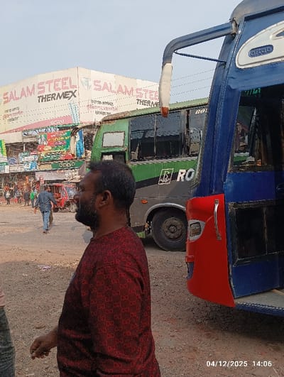 An outdoor scene in Kaliganj, Bangladesh, is depicted on April 12, 2025, at 14:06. The image features a man in the foreground, positioned right of center, wearing a dark red, long-sleeved shirt with a repetitive pattern. He has a beard and is looking towards the left, with his right hand extended downwards. To the immediate right of the man is the front-right section of a blue and red bus, partially visible. The passenger door is open, and a rearview mirror extends from the frame. The word "AFTAB" is discernable on the blue portion above the windshield.
Behind the foreground subject and the blue bus, a green bus with "ROYAL" branding on its side is parked. Its windows are tinted dark. In the background, a busy street or market area is visible. A large white sign reading "SALAM STEEL THERMEX" is prominently displayed on a building. Below this sign, numerous smaller storefronts and advertisements, some in Bengali and one with "OPPO" branding, line the street. Several individuals are present in the background, including one person in a blue shirt walking away from the camera. The ground is unpaved, consisting of dirt and gravel. The sky overhead is clear.