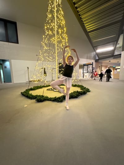 A young ballerina strikes a pose in front of a Christmas tree illuminated with warm white lights. She wears a black leotard and pale pink tights with matching ballet shoes. A wreath encircles a circular object on the floor near her. Other people are visible in the background, along with informational kiosks. The setting appears to be a public indoor space with a high, corrugated metal ceiling.