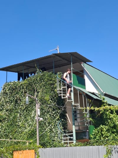 In the village of Shchebetovka, Ukraine, a man stands on an outdoor spiral staircase, descending from a building covered in dense green ivy. The man, wearing a striped tank top and dark shorts, carries a backpack and appears to be holding a cigarette. The staircase is made of metal with a light green railing. The building has a corrugated metal roof and green siding. A street lamp with a glass fixture is visible in the foreground, attached to a utility pole. A grey metal fence runs along the bottom of the image, with a section of wooden fencing visible to the left. The sky is clear and blue.