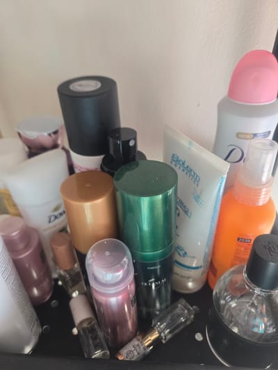 A collection of assorted cosmetic and personal care products is arranged on a dark surface, likely a shelf or tray. Among the visible items are several bottles and tubes of varying sizes and colors. A prominent white tube with blue lettering, possibly a face cream, stands towards the center. To its right, an orange spray bottle is visible. A large black-capped bottle is positioned behind a metallic gold-colored cap. Other items include a pink-capped white bottle, smaller spray bottles, and what appears to be a men's fragrance bottle with a clear glass body and a black cap. The background is a plain, light-colored wall. The scene is illuminated with artificial light, casting shadows on the objects. This arrangement suggests a personal vanity or storage area. The location is identified as Deira, United Arab Emirates.