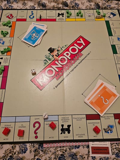 This is a top-down shot of a Monopoly board game set on a patterned fabric surface. The game appears to be in progress, with several tokens and plastic houses scattered across the board.
The subjects are the Monopoly board itself, various game pieces, and cards. Visible objects include a silver car token, red houses, green houses, two dice (one showing a '3' and another showing a '1'), and several Monopoly property cards and Chance cards.
The scene is a board game being played. The activity taking place is implied by the placement of tokens and houses, suggesting players are moving their pieces and developing properties.
Notable details include the prominently displayed "MONOPOLY" logo on the board, as well as various property names and monetary values printed in Russian. Some text visible includes property names like "НАГАТИНСКАЯ УЛ." (Nagatin Street), "ШАНС" (Chance), "ОБЩЕСТВЕННАЯ КАЗНА" (Public Treasury), and monetary values such as "M100", "M200", and "M120". The fabric underneath the board has a blue floral pattern. There is no indication of the time of day or weather, nor any specific location cues beyond the Russian text on the board. The overall mood is neutral, focused on the game.