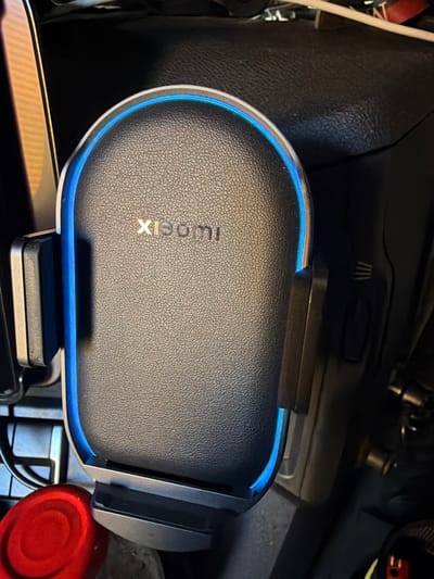 The image shows a close-up, top-down view of a black car phone mount with a blue LED light. The phone mount has a textured, leather-like surface and the "XIAOMI" logo is visible in gold lettering on the front. The mount is clipped onto what appears to be a car's center console. There are other objects visible in the background, including a red circular object, some wires, and dark gray plastic surfaces, suggesting the car interior. The lighting is dim, with the blue light of the phone mount being a prominent light source. The image's context does not provide any information about the city of Arques, France.