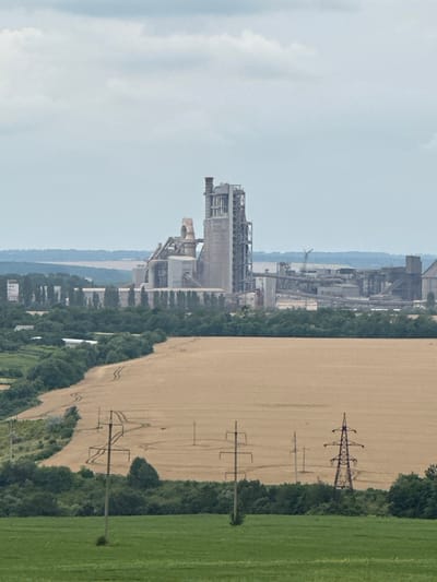 The image is a wide-angle, eye-level outdoor photograph taken on an overcast day, looking towards a large industrial complex. In the foreground, a vibrant green field transitions to a harvested wheat field, with tire tracks visible in the dry stalks. Several electrical poles with wires crisscross the landscape. The midground features a dense line of trees separating the agricultural fields from the industrial zone. The background is dominated by a sprawling factory with tall, concrete structures, silos, and various interconnected pipes and buildings, indicative of a cement plant or similar heavy industry. A crane is visible within the industrial complex. The sky is a muted gray with scattered clouds. The scene appears to be located in the vicinity of Kolubaivtsi, Ukraine, a region known for its industrial sites. No people are visible in the image.
