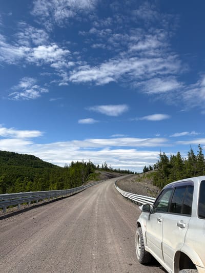In Anavgai, Russia, a silver SUV is parked on a dusty gravel road. The vehicle is positioned on the right side of the frame, with its side facing the viewer. The road curves to the left, disappearing into the distance. On the left side of the road, a dense, green forest covers a hillside. On the right side of the road, the terrain is rocky and sparse, with scattered evergreen trees. The sky above is a vibrant blue, with scattered white clouds. The scene depicts a remote, natural landscape.