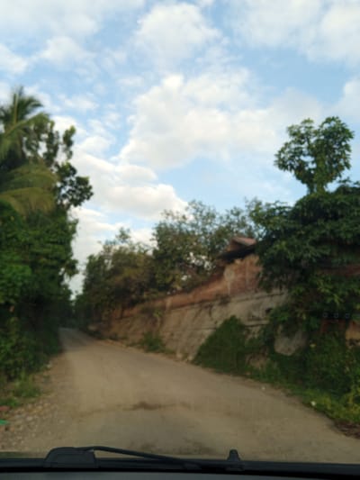 The image depicts a dirt road in Awegeutahpaya, Indonesia. Lush green vegetation lines both sides of the road, with trees partially obscuring a dilapidated structure on the right. The sky is blue with scattered white clouds. The view is from inside a vehicle, evidenced by a dashboard and windshield wipers at the bottom. There are no people visible.