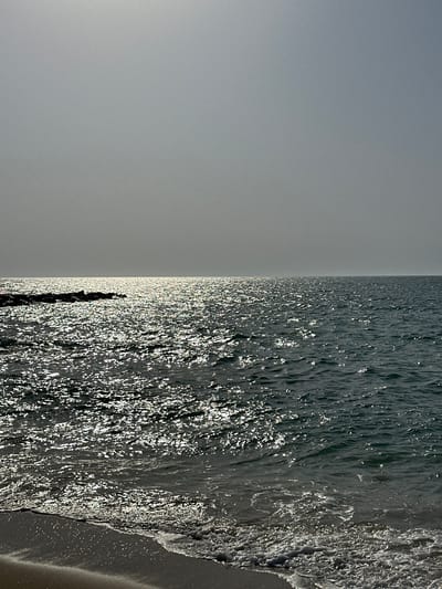 The image shows a wide shot of the sea under a hazy sky. The sun reflects off the choppy water, creating a shimmering effect. On the left side of the image, a rocky outcrop juts out into the sea. In the foreground, the edge of a sandy beach is visible with small waves lapping at the shore. The overall impression is a serene, overcast day at the coast. The location is specified as Al Jazeerah Heritage Village in the United Arab Emirates. There are no people or specific objects of interest apart from the natural elements of the sea, sky, and rocks.