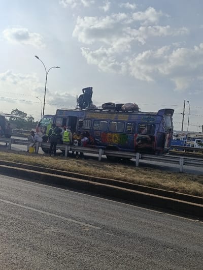 The image depicts a purple public service minibus (matatu) stopped on the shoulder of a multi-lane road in Kiuu ward, Kenya, under a partly cloudy sky. The matatu is heavily customized with colorful graffiti, prominently displaying "OCCUPY" on its side. Its roof rack is loaded with multiple spare tires and a white sack, among other items. One male individual, dressed in dark clothing, is visible standing on the roof rack, actively engaging with the cargo. Below, approximately seven to ten individuals are gathered around the front and side of the matatu; at least two are wearing high-visibility vests. A person near the front of the vehicle is holding a white bucket. The matatu is positioned beside a metal guardrail separating it from a grassy median strip. A paved road, marked with a white lane line, occupies the foreground. A street light pole is visible to the left of the matatu, with additional poles and distant vehicles in the background. The scene suggests a temporary roadside stop, potentially for loading, unloading, or addressing a vehicle issue.