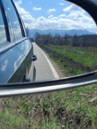 The image shows a view from a car's side mirror. The road stretches out into the distance, flanked by green foliage on either side. A mountain range is visible in the background, partially obscured by clouds. The sky is blue with scattered clouds, which are reflected on the surface of the car. The location is Komarevo, Bulgaria.