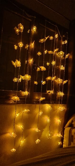 The image depicts an indoor scene in El Campello, Spain, illuminated primarily by decorative string lights. Multiple vertical strands of amber-yellow, translucent, flower-shaped or butterfly-shaped LED lights are suspended, forming a luminous curtain. These lights are positioned in front of a dark, multi-paned wooden window or door frame, with their reflections visible on the glass. A dark, possibly metallic, fixture resembling a handle is discernible on the frame. The ambient light originates solely from these luminaires, casting a soft, warm glow on a textured wall surface visible to the left and below the frame. The foreground features an indistinct horizontal surface, consistent with furniture covered in dark fabric. A light-colored object with vertical stripe patterns, possibly a pillow or bedding, is partially visible on the far right of the scene. No individuals are present, and no specific actions or interactions are depicted.