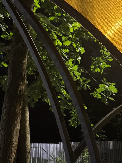 The image depicts an outdoor scene at night, possibly in Doma 1096 Km, Russia. The foreground features two tree trunks with textured bark. Behind the trees, several green oak leaves are illuminated, suggesting artificial lighting. Overlapping the scene are dark, curved metal beams. In the upper right corner, a yellow, textured material is visible, possibly a canopy or awning, which is also illuminated. In the background, a vertical metal fence is discernible. The overall impression is of an outdoor structure with natural elements at night. There are no people or animals present.