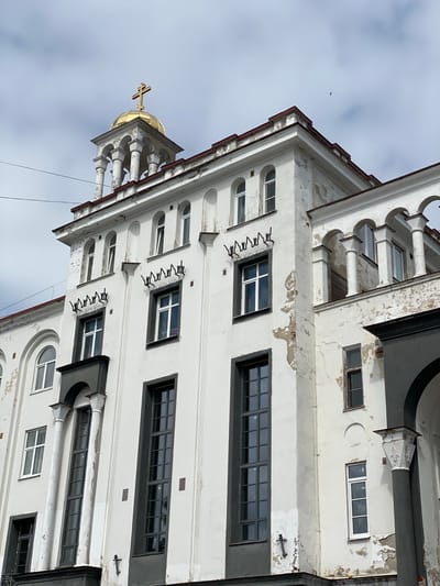 The image shows a white building with peeling paint, featuring a golden dome topped with a cross. The building has numerous windows of various sizes and styles, some with dark frames and others with white frames. Decorative elements, such as columns and arched openings, are visible. There are also some metal sculptures attached to the facade of the building. The sky is cloudy and overcast. The building is located in Sortavala, Russia.