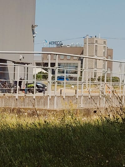 The image depicts an outdoor scene on a sunny day in Łomna-Las, Poland. In the foreground, a patch of overgrown green grass and weeds obscures a concrete barrier. Behind this barrier, a white metal railing runs horizontally across the frame, separating the foreground from the middle ground.
In the middle ground, there is a road or pathway. Parked on this pathway are two bicycles and a green and black scooter. Further down the pathway, a dark car is visible, and beyond that, a blue vehicle is driving. The background features a modern, multi-story building with a light-colored brick facade and large windows. A sign on top of part of the building clearly displays the text "AERECO" in blue capital letters, with a stylized blue logo above it. The sky is a clear, light blue, suggesting fair weather. The overall impression is one of a contemporary urban or industrial setting bathed in bright daylight. There are no people visible in the image.