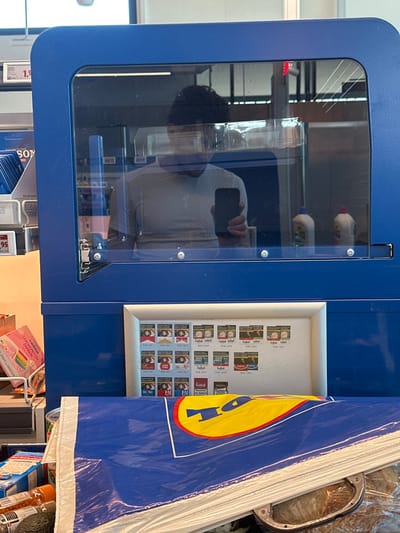 The scene depicts the checkout area inside a Lidl supermarket located in Jestetten, Germany. The viewpoint is from a first-person perspective, looking towards the cashier's station.
In the foreground, a stack of blue plastic shopping bags with the yellow "Lidl" logo is prominently displayed, partially obscuring groceries such as an avocado and other packaged items on what appears to be a conveyor belt or bagging area.
Above the shopping bags, a blue structure, likely part of the checkout counter, features a clear, transparent protective screen secured with multiple metal fasteners. Reflected within this screen is the upper torso and head of a person with dark, curly hair, wearing a light-colored t-shirt. This individual is holding a black smartphone with their right hand, indicating they are taking the photograph. The reflection also shows parts of the supermarket interior, including fluorescent lighting fixtures and two white plastic bottles with colored caps.
Below the protective screen, integrated into the blue counter structure, is a framed display showcasing various tobacco products, including cigarettes from brands such as Marlboro, Lucky Strike, Goldfield, Roth-Händle, EM, HB, and Bodega. Each product image includes German text warning labels and pricing (e.g., "20 St. / 7,20 €"). To the left of this display, a rack containing rolling papers, with a "SONY" branded package partially visible, displays price tags in Euros (e.g., "1,99" and "1,95"). A pink item, possibly a flyer or magazine, is also present on the far left.