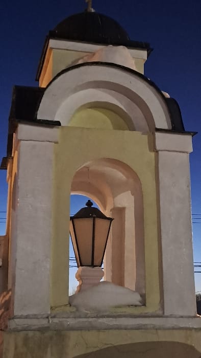 The image depicts a section of a light-yellow and off-white architectural structure, identified as being in Avdot’ino, Russia, under a deep blue evening or night sky. The structure features a dark, dome-shaped roof topped with a small, golden Orthodox cross. Below the dome, snow accumulates on ledges and architectural elements. The main visible section comprises arched openings supported by vertical columns, painted in alternating light-yellow and off-white tones. Centered within one of these arches is a large, dark-framed lantern with illuminated, light-colored panels, resting on a decorative, off-white pedestal. Snow is visible at the base of the lantern and on the pedestal. Cracks are present in the lower sections of the plaster or paint. Faint dark lines, possibly utility cables, are visible against the sky in the background to the right of the lantern. No individuals are present.