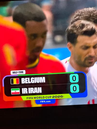 This is a close-up, eye-level, medium shot of a television screen displaying a soccer match scoreboard. The screen shows "HALF-TIME" in a white rectangular box on the left. Below this, the scoreboard lists two teams: "BELGIUM" with its flag and "IR IRAN" with its flag. Both teams have a score of "0" displayed next to them in turquoise boxes. At the bottom of the scoreboard, it reads "FIFA WORLD CUP 2026" in a neon green and blue design, with "FIFA.com" in a white button. The background of the image consists of blurry figures of soccer players. The image is not in the city of Bykovo, Russia.