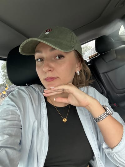 A young woman with light brown hair, wearing a green baseball cap with a mushroom embroidered on it, and a light blue button-down shirt over a black top, rests her head on a black car seat. She has on a silver watch with a link band on her left wrist and a gold necklace with a clover pendant. Her right hand is resting near her face, and she is wearing rings and has white-tipped nails. The woman has brown eyes, defined eyebrows, and light pink lipstick. Her expression appears neutral or slightly bored.
The scene is inside a car, as evidenced by the car seats and the window in the background. Through the window, it appears to be raining, with water droplets visible on the glass. The weather seems overcast and possibly dull. The interior of the car is dark, with black leather seats. A sticker on the back of the headrest is partially visible and contains the text: "DO NOT FOLLOW." The lighting suggests it is daytime, but the overcast weather makes it appear dim. The woman is likely a passenger in the car, possibly on a road trip or just commuting. The overall mood of the image is casual and relaxed, with a hint of a melancholic or introspective feeling due to the weather and her expression. The location context provided is Tangelo Park, USA.
