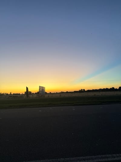 A photo capturing a vivid sunset next to a road. The sky transitions from blue to golden hues. A vertical beam of light cuts through the sky. A billboard is visible, silhouetted against the setting sun. Trees line the horizon. The road, occupying a significant portion of the image, is dark and textured.