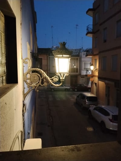 An exterior, low-angle, eye-level shot captures a street scene in the city of Sueca, Spain, at dusk. The image is dominated by an ornate, vintage streetlamp bracket extending from a light blue building wall on the left. The bracket is white and features intricate scrollwork and a circular emblem. The illuminated streetlamp itself, with its crown-like top, is a prominent element on the right side of the bracket.
The street below is paved and appears to be a narrow European-style street, lined with buildings on both sides. Several cars are parked along the street, including a white SUV, a silver van, and a dark red hatchback. A red and white STOP sign is visible on the right side of the street. The sky above is a deep twilight blue, with visible antennas on some of the rooftops. The buildings are multi-story structures with windows and balconies. The left building is painted light blue, while the buildings on the right are an ochre or light brown color. The scene is devoid of people. The overall impression is of a quiet, urban street at the end of the day.