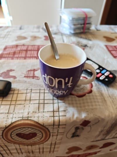 On a patterned tablecloth, a purple mug with "don't worry" printed on it sits beside a black remote control. A silver spoon is inside the mug, which contains a light brown beverage. In the background, there is a stack of items secured with a pink rubber band. The location appears to be an indoor setting, potentially a home, based on the presence of common household items. The specific location within Heraklion Municipal Unit, Greece, cannot be determined from the image.