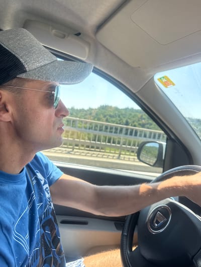 A close-up, low-angle shot shows a man driving a car. The man is wearing a grey baseball cap and sunglasses, and a blue t-shirt with a white graphic design. His right arm is on the steering wheel, which has the Dacia logo in the center. The car's interior is visible, including the sun visor and the window. Outside the car, trees and a bridge are visible. The sun is shining, suggesting it is daytime. The man appears relaxed. The image has a vignette effect, with the edges darkened. There is a yellow sticker with some text and images on the top right of the windshield. The crops show some detail of the car interior, the side mirror reflecting the outside, and the man's arm and t-shirt. The location is identified as Villards-d'Héria, France.