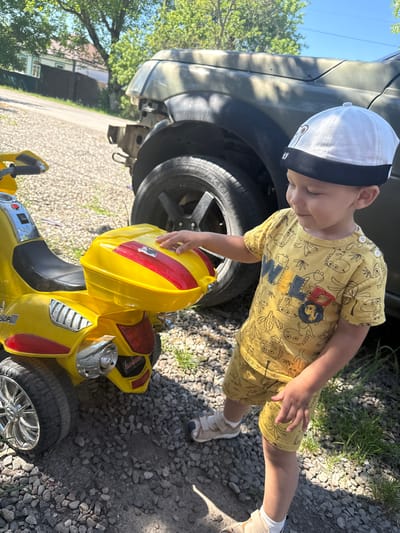 A young boy wearing a yellow t-shirt with a dinosaur print and a white cap stands outdoors in Vinsady, Russia, near a yellow toy motorcycle and a damaged olive-green SUV. The boy is reaching out to touch the back of the toy motorcycle, which has a yellow storage box. The ground is covered in gravel and some patches of grass. The SUV appears to have a damaged front bumper and headlight area, with its camouflage paint faded. In the background, there are trees and a building with white siding and a dark roof, possibly a residential house. The sky is clear and blue, indicating a sunny day.
