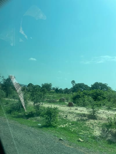 A low-angle, medium shot captures a scenic view of a rural landscape in Mararaban Tula, Nigeria, seen through a vehicle's window. The sky, a vibrant turquoise, occupies the upper half of the frame, with a few wispy white clouds scattered across it. A reflection of the interior of a vehicle, including what appears to be a seat and a headrest, is visible on the left side of the glass.

The landscape below is a mix of green vegetation and sandy terrain. Lush green grass covers the foreground, transitioning to patches of dry, sandy earth interspersed with shrubs and trees. Several large, rounded rocks are scattered across the middle ground. A prominent palm tree stands out among the denser foliage in the distance, towards the right of the frame. The overall impression is one of a dry, sun-drenched environment with sporadic bursts of greenery. The image quality is slightly degraded, possibly due to the window glass or a low-resolution capture.