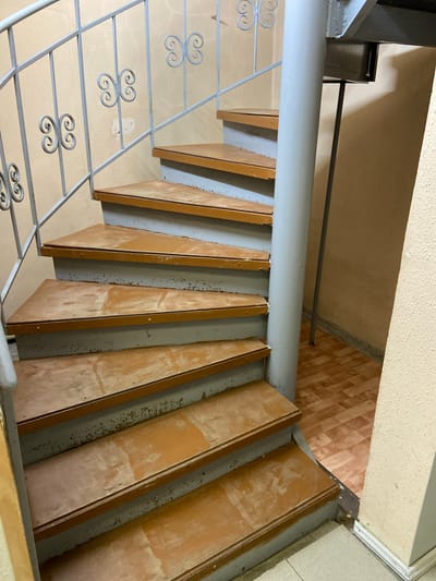 The image shows a close-up, eye-level view of a spiral staircase with wooden steps and a grey metal railing. The steps are a light brown color with some wear and tear, and the risers are grey. The railing is made of thin metal bars with decorative butterfly-shaped elements. A thick, grey cylindrical pole is located to the right of the stairs, likely serving as a central support. The walls appear to be painted a pale yellow or cream color. The flooring visible at the bottom of the stairs is a wood-look linoleum with a pattern of light and dark brown rectangles. The overall impression is of an indoor, utilitarian space, possibly in an older building. There are no people or animals visible in the image, and no activities are taking place. The scene is static. The location is identified as Kraskino, Russia, though no specific landmarks or context within the city are discernible from the image alone.