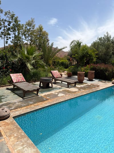 This image captures a serene outdoor setting, likely a poolside area at a resort or private villa in Tahannaout, Morocco. The scene is bathed in bright daylight under a clear blue sky, accented by wispy white clouds.
The most prominent feature is a rectangular swimming pool, occupying the right side of the frame. Its vibrant blue mosaic tiles are clearly visible beneath the clean, still water, with a few small leaves floating on the surface. The pool is bordered by a rustic, weathered coping made of concrete or stone in earthy tones.
Along the left edge of the pool, a paved patio area features several wooden lounge chairs, each topped with colorful, patterned cushions, suggesting comfort. A small wooden barrel-shaped table is placed between some of the chairs. Further back on the patio, large, decorative terracotta pots add to the natural aesthetic. The strong sunlight casts crisp shadows of the chairs and pots across the patio.
Beyond the patio, the landscape is lush with diverse greenery. Various trees with dense foliage, including prominent palm trees and flowering bushes with hints of red blossoms, create a verdant backdrop. In the distant background, under the expansive sky, a majestic, rocky mountain range is visible, typical of the Atlas Mountains region surrounding Tahannaout.
The overall impression is one of peace and tranquility, inviting relaxation in a warm, sunny environment. There are no people visible, and no discernible text is present in the image.