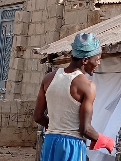 In Gayawa, Nigeria, an adult male stands in profile, facing right. He wears a light-colored sleeveless tank top, blue shorts with a white stripe, and a teal patterned headwrap. His left hand, visible at the lower right, is covered by a bright red glove. His mouth is slightly ajar. Behind him, a corrugated metal roof is visible, along with white fabric, possibly a canopy or curtain, to the right. To the left, a building constructed of light-colored bricks features a window with a dark metal security griller. The ground consists of compacted earth.