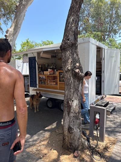 A woman lies on a large tree trunk in front of a white food truck. A man with bare chest stands in the foreground holding a phone. A dog stands beside the truck. Electrical equipment sits next to the tree. The scene is outdoors on a sunny day with trees and foliage visible.