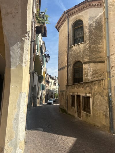 The image depicts a narrow cobblestone street in Maser, Italy, flanked by aged buildings. On the left, a pale yellow building with peeling plaster dominates the foreground, featuring arched openings. To the right, a beige, weathered building with a curved facade showcases arched windows with intricate metal grilles. The street continues into the distance, lined with more buildings displaying architectural details such as balconies with greenery and awnings. Sunlight casts shadows across the cobblestones, indicating a clear day. A white car is parked on the street in the mid-ground, and a dark car is visible further down the street. The sky is blue with scattered clouds.