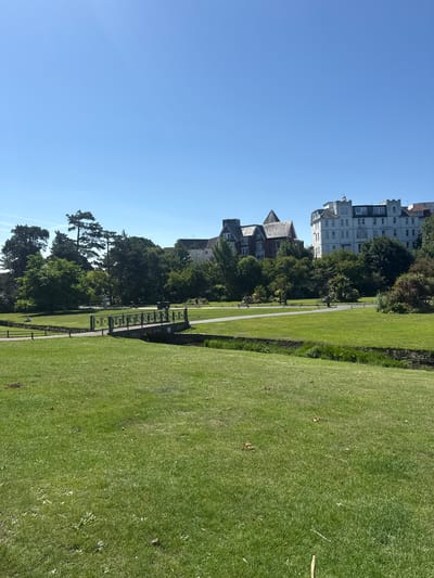 The image depicts a sunny day in Bournemouth's Lower Gardens, a public park in Bournemouth, United Kingdom. In the foreground, a lush green lawn slopes gently upwards. A narrow, stone-arched bridge with ornate metal railings crosses a small stream or ditch that bisects the landscape. Beyond the bridge, a paved pathway winds through the park, bordered by more manicured lawns and mature trees. In the mid-ground, several people are visible: one individual sits on the grass near the bridge, while others walk along the pathway. In the background, large, multi-story buildings with white facades and traditional architecture are visible, suggesting the proximity of the town center to the park. The sky is a clear, bright blue, indicating pleasant weather. The overall scene conveys a peaceful and well-maintained urban park setting.