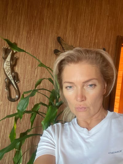 The media file shows a close-up, sideways shot of a fair-skinned woman with blonde hair, blue eyes, and light makeup. She is wearing a white t-shirt with the brand name "DEMIX" visible on the left side of her chest. The woman is positioned in the upper right portion of the frame.
The setting appears to be indoors, with a textured wooden surface dominating the lower and left sides of the image. A long, trailing green plant with numerous leaves is draped across the wooden surface, extending from the right side of the frame towards the bottom left.
In the bottom left quadrant of the image, there is a decorative wooden carving of a lizard or gecko, intricately detailed with inlaid patterns on its back and sides. The lighting suggests it is daytime, possibly indoors with ambient light, as there are no strong shadows. The overall mood is casual and personal, likely a selfie. No specific activity or event is depicted beyond the pose of the individual.
The location context provided is Габишево, Russia.