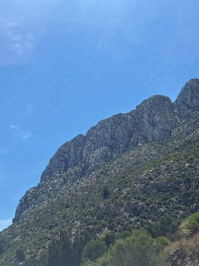 The image shows a mountainside in El Verger, Spain, viewed from a lower angle. The mountain's rock face is visible, with varying shades of gray and green vegetation clinging to its surface. The sky is a clear, bright blue with a few wispy clouds.