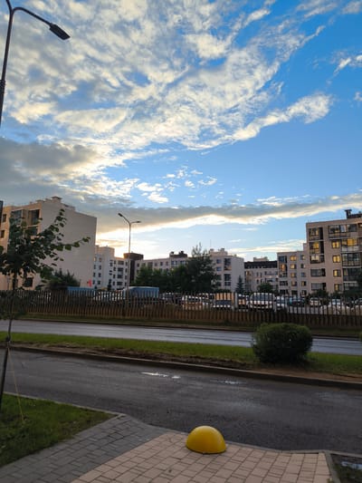 This image depicts a street-level view of a residential area in Otradnoye, Russia. The scene is dominated by several modern apartment buildings, characterized by their clean lines and light-colored facades. Some buildings have balconies and large windows, with lights visible in some of the apartments, suggesting it is either early morning or late evening.
In the foreground, a paved sidewalk with a distinct brick pattern leads the eye towards a bright yellow, dome-shaped bollard. Adjacent to the sidewalk is a strip of green grass, followed by a wet asphalt road. The road appears to have puddles, indicating recent rain. A wooden fence runs along the far side of the road, separating it from a grassy area and parked vehicles. Various cars and vans are visible behind the fence, suggesting a parking area for residents.
The sky above is a prominent feature, with a mixture of blue sky and scattered, wispy clouds. The clouds are illuminated by what appears to be the setting or rising sun, casting a warm, golden glow on some of them, while others are tinged with gray. A street lamp stands tall on the left side of the image, silhouetted against the sky. Another street lamp is visible further down the road, between the apartment buildings. Trees and bushes are scattered throughout the scene, adding touches of green to the urban landscape.
The overall mood of the image is serene and tranquil, amplified by the soft lighting and the quiet atmosphere of the residential setting. There is no visible text in the image.