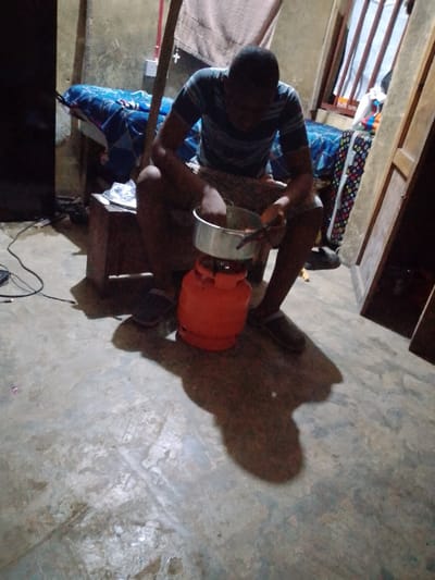 An individual is seated, stirring a metal pot on a small, orange gas tank. The scene is indoors, with a wooden table and a bed visible in the background. The person is wearing a striped shirt and shorts. The lighting is dim, creating distinct shadows on the floor. The setting appears to be a modest living space.