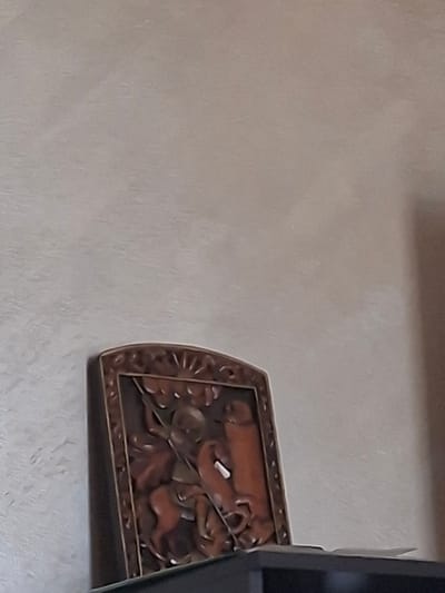 A carved wooden plaque depicting a warrior on horseback is displayed on a shelf. The plaque is rectangular with a decorative border. The carving shows a figure in armor with a sword, facing a dragon or serpent-like creature. The warrior is mounted on a horse that is rearing. The background of the plaque is textured. The plaque is positioned on the right side of a dark shelf, with a portion of the shelf and the wall behind it visible. The wall has a light, textured finish. The location in Aksakovo, Bulgaria is not discernible from the image.