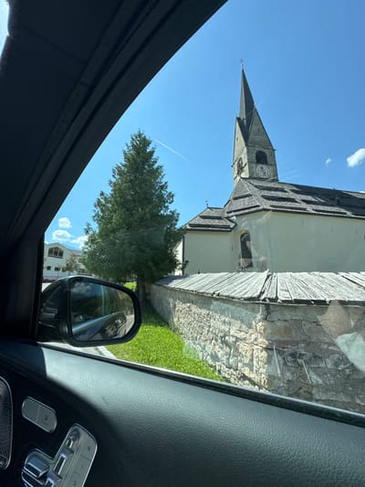 The image, captured from inside a vehicle, shows a view through the side window of a church in San Martin de Tor - San Martino in Badia - St. Martin in Thurn, Italy. The church features a tall, pointed steeple with a clock face and a dark tiled roof. A stone wall with a weathered wooden fence runs in front of the church, with a well-maintained grassy area behind it. A large, dark green conifer tree stands to the left of the church. The sky is clear blue with a few white clouds and a faint contrail. The interior of the car, including part of the door panel and window controls, is visible in the foreground. The side-view mirror reflects the road and other vehicles.