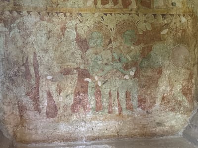 The image depicts a deteriorated ancient fresco painting on a wall, likely from a cave temple. The painting appears to show a group of figures, possibly deities or royalty, adorned with headwear and garments. Two figures in the center seem to be holding musical instruments, possibly stringed. Foliage or floral designs fill the upper portion of the painting. The location is likely a cave temple within the ancient city of Polonnaruwa, Sri Lanka. The painting is faded and damaged by the effects of age and the elements.