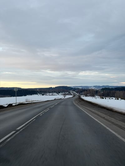 The image depicts a two-lane asphalt road, likely located in Vakhrino, Russia, viewed from a moving vehicle. The road features a solid white line marking the right shoulder and a dashed white line separating the two directions of travel, curving gently to the left in the foreground and more sharply into the distance. Patches of residual snow, mixed with dirt and mud, line both sides of the road, indicating thawing conditions. Beyond the immediate roadside, fields covered in melting snow extend, interspersed with bare deciduous trees and shrubs. In the mid-ground and background, dense evergreen forests cover undulating terrain, with a distant settlement visible among the trees and a prominent cylindrical water tower on a hill. A circular road sign is partially obscured on the left shoulder. The sky is uniformly overcast with grey clouds, showing a faint light yellow tint near the horizon. No individuals or active subjects are visible within the frame. The overall environment suggests a late winter or early spring day.
