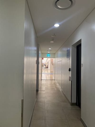This image captures a brightly lit, narrow indoor hallway within a commercial or public building located in Gangnam-gu, South Korea. The perspective is directed down the length of the corridor, which features light-colored, square-tiled flooring and plain, light-toned walls with a slight sheen reflecting vertical light streaks. The white ceiling is equipped with multiple recessed circular light fixtures, a prominent circular air vent/diffuser in the foreground, and smaller circular sensors or smoke detectors.
On the right wall, a dark, closed door is set within a dark frame. Above and slightly to the left of this door, a rectangular sign with unidentifiable text and a symbol is mounted. Adjacent to the door, a black wall-mounted access control device, possibly a card reader, and a red wall-mounted fire safety device, likely a pull station, are present.
At the far end of the hallway, a wide doorway opens into a spacious, highly illuminated public area with reflective polished floors. A green, illuminated exit sign displaying a running human pictogram is centrally positioned above this doorway. Beyond the doorway, several indistinct human figures are visible, indicating activity within the brightly lit space, which appears consistent with a shopping mall or large facility lobby. No individuals are present within the immediate hallway.