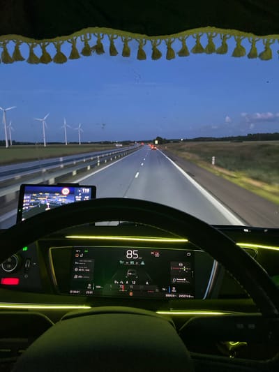 The image is taken from the perspective of a person driving a truck at dusk or night on a highway. The dashboard of the truck is illuminated with green LED lighting. A digital display shows the speed as 85 km/h, along with other driving information such as fuel consumption and distance traveled. There is also a GPS navigation screen displaying a map. Outside the truck, a highway stretches into the distance, with streetlights and other vehicles visible. Wind turbines are lined up along the side of the road, indicating the truck is likely driving through a rural or open area. The sky is dark blue, suggesting it is either early morning or late evening. The image is stated to be located in the city of Långås, Sweden.