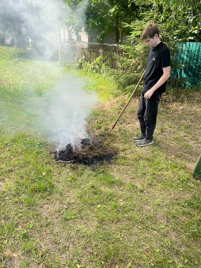 The image shows a young person wearing a black t-shirt and black pants, standing in a grassy outdoor area. They are looking down and appear to be tending to a small fire with a pitchfork. Smoke is rising from the fire. In the background, there is a green fence, trees, and a brick building. The scene suggests a sunny day, likely in a rural or suburban setting. The person's posture and gaze might indicate concentration on the task at hand. There is no visible text in the image. The context provided indicates the location as Babinitski rural council, Belarus.