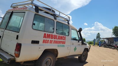 This outdoor scene captures a white, rugged 4x4 ambulance, likely a Toyota Land Cruiser model, parked on a dirt road under a bright blue sky with large, fluffy white clouds. The ambulance, facing towards the right and slightly away from the viewer, is equipped with a roof rack and a snorkel, indicating its suitability for challenging terrain.

Red lettering on the side clearly spells "AMBULANCE" and "HGR DE RWAMPARA". Below this, two green phone numbers are visible: "+243 810119669" and "+243 827363209". A prominent green cross symbol is painted on the front passenger door. The vehicle appears somewhat dusty, particularly on its lower body and tires, suggesting recent travel on unpaved roads.

In the background, a dirt road stretches further, where several motorbikes are parked. A few people are standing near the motorbikes and simple structures, possibly stalls or buildings, are visible. Green trees and distant mountains complete the horizon. The bright sunlight suggests it is midday or early afternoon, with clear weather. The overall setting points to a rural or semi-urban area in Tchere, Democratic Republic of the Congo, consistent with the local context and the "HGR" (likely Hôpital Général de Référence) designation. A watermark "Joseph Kisuki" is present in the bottom right corner.