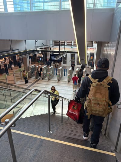 The image depicts a bustling, modern transit station entrance, likely part of a metro or light rail system in Brossard, Canada. The vantage point is from the top of a set of dark grey granite stairs, looking down towards the station concourse and multiple rows of turnstiles.
In the foreground, a person is seen from behind, wearing a black cap, black hoodie, black pants, and white and black sneakers. They are carrying a large tan/khaki tactical-style backpack and holding a red shopping bag that clearly displays the word "metro". The stairs on the right feature a bright yellow safety line and metal handrails. Further down the stairs, another individual in a yellow-green jacket and sunglasses is visible, leaning against a railing and carrying a black bag.
The midground is dominated by sleek, automated turnstiles, some showing green arrows indicating entry and others red crosses for closure. Several commuters are actively passing through these turnstiles, including one person in a red shirt carrying a blue bag. Above the turnstiles, digital display screens show information, though the text is unreadable.
To the left, a prominent, linear light fixture runs vertically, illuminating the area. Large windows with horizontal grates are visible on the far left, allowing natural light into the station. The overhead structures consist of clean, white panels, contributing to the station's contemporary design.
The scene captures typical daytime commuter activity within a clean and well-lit public transport hub. A directional sign in the background includes the text "Avenue Tisserand" and "14 RTC", likely indicating a street name and possibly a bus line or route number. The overall atmosphere is one of routine movement and transit.