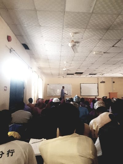 The image displays the interior of a lecture hall or large classroom located in Panhauya, Nigeria. A male individual, identified as an instructor, stands at the front of the room, facing an audience. He is dressed in a light-colored, long-sleeved garment and a cap, with his right arm extended and gesturing towards a whiteboard mounted on the left wall. A second, blank whiteboard is mounted on the right side of the front wall, alongside a wooden podium and a projector stand.
Approximately 30 to 50 individuals are seated in rows, facing the instructor, with their backs predominantly visible to the viewer. Several attendees wear distinct head coverings, including white and patterned caps. Some individuals have notebooks or papers open on a surface in front of them.
The room features light-colored walls and a white grid panel ceiling from which three white ceiling fans are suspended. Several ceiling panels are dislodged or missing. A red circular fire alarm is mounted high on the upper left wall, beneath a linear light fixture. A bright light source, likely a window or open doorway, is present on the left side of the room, causing localized overexposure.