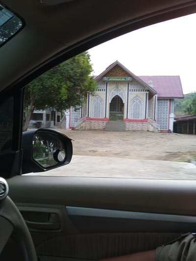The image shows a view from inside a car looking out the driver's side window at a mosque. The mosque is a light-colored building with a red roof and intricate white latticework on its facade. There are stairs leading up to the entrance, and trees are visible behind the mosque. The car's interior is visible in the foreground, including the steering wheel and a side mirror. The image is taken from a fixed perspective, suggesting it was captured while the car was stationary or moving slowly. The environment appears to be a rural or semi-rural setting in Luengbaro, Indonesia, given the style of the mosque and the surrounding vegetation. There are no people clearly visible in the scene.