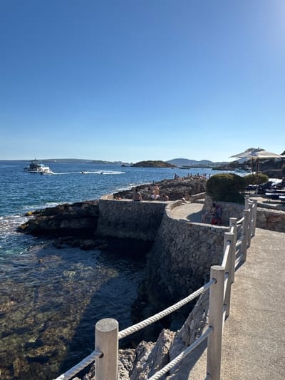 The image depicts a coastal scene in Cas Català-ses Illetes, Spain, under a clear blue sky. In the foreground, a paved walkway with a wooden railing and rope accents curves along a rocky coastline. The sea, a deep blue, laps against the rocks, revealing clear water with visible seabed. Further out, a white passenger boat navigates the water, followed by a jet ski. Along the rocky outcrop, several individuals are scattered, some sunbathing on the rocks, while others are seated on chairs or the stone wall. On the walkway, more people are visible, with some seated in chairs and one individual standing near a large sun umbrella. In the background, the coastline stretches with hills and buildings. The overall atmosphere suggests a sunny day at a seaside resort or public area.