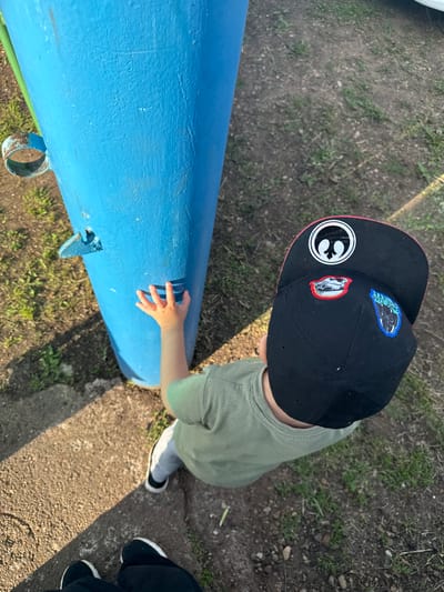A young child wearing a green t-shirt and a black baseball cap with patches is reaching out to touch a bright blue metal pole in an outdoor setting. The child's left hand is extended, fingers spread, towards the pole. The ground around the pole is a mix of dirt and sparse grass. In the foreground, two pairs of feet are visible; one pair belongs to the child, wearing black and white sneakers, and the other pair, in black shoes, suggests an adult is present, possibly taking the photo. The lighting indicates it is daytime, with shadows cast across the ground. The location is identified as Чуй-Атасево, Russia.